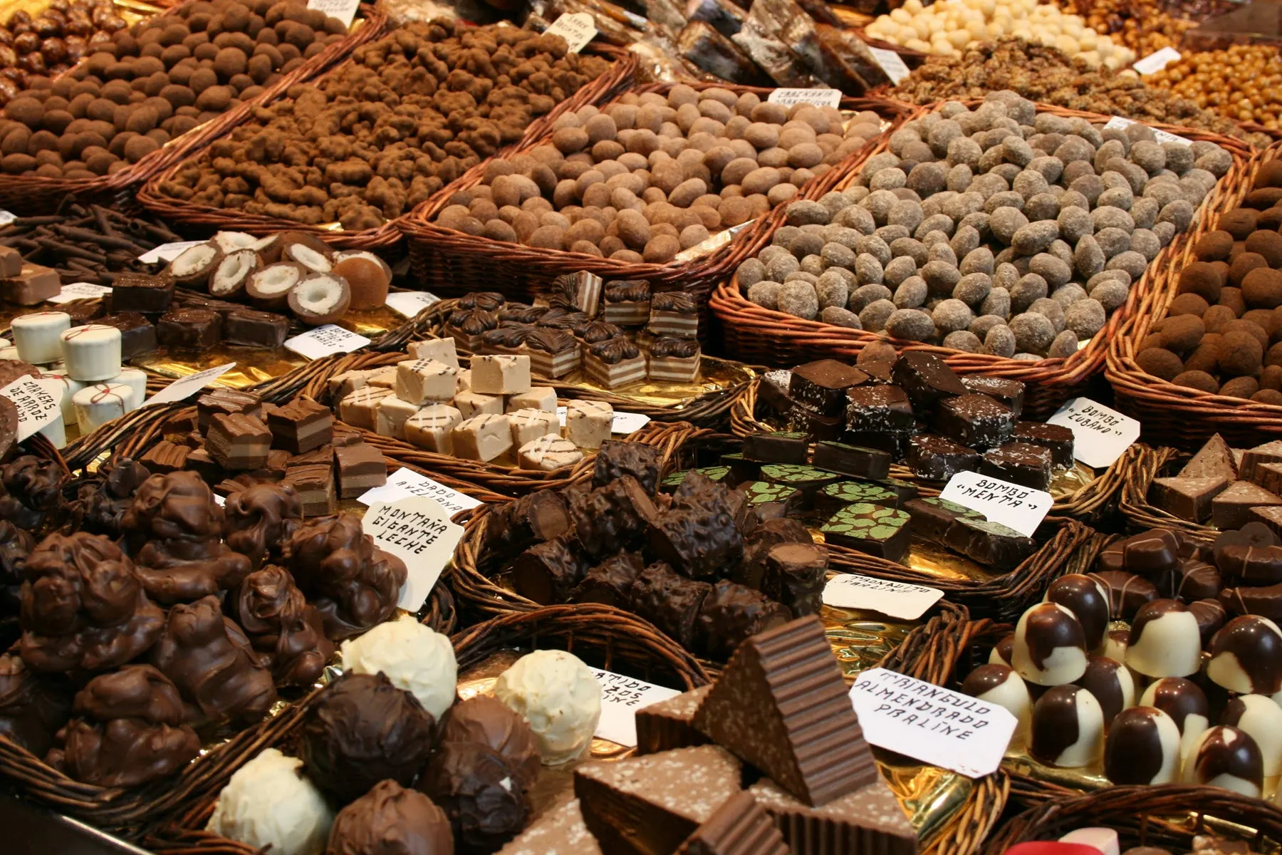 Chokladpraliner och tryfflar i flätade korgar på ett marknadsstånd, varje sort märkt med en handskriven namnskylt.